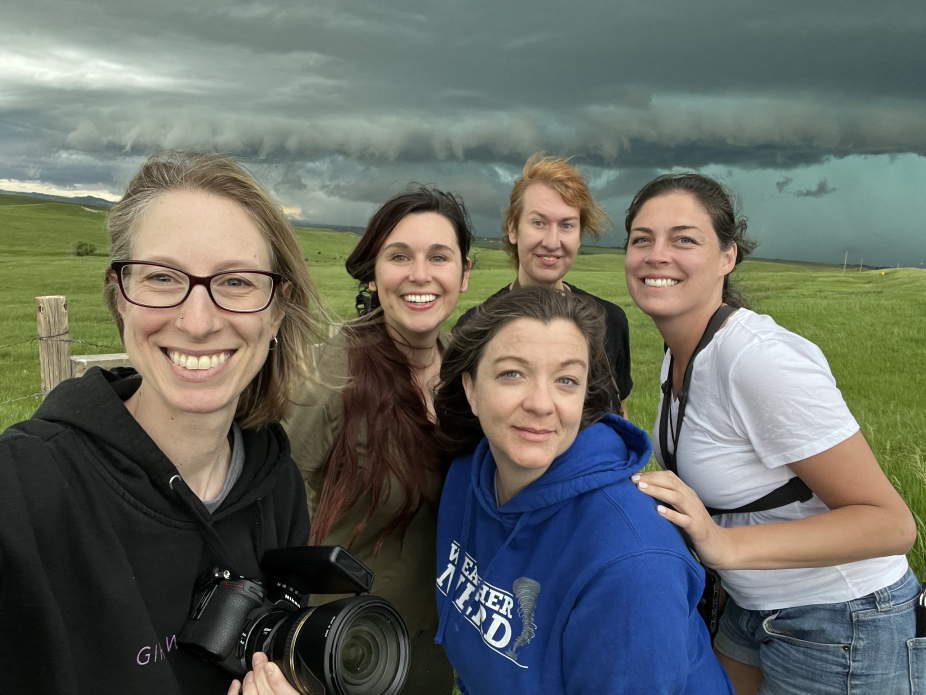 Five women storm chasers pose in front of an incoming storm. 